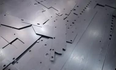 Close-up of flat, laser-cut grey sheet metal parts laid out flat on a surface. The pieces have precision geometric cutouts, slots, and circular holes ready for industrial assembly or bending.