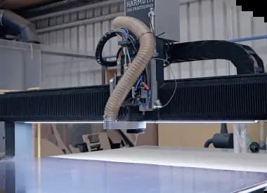 A close-up shot of an industrial CNC milling or cutting machine inside a workshop. The machine features a large black horizontal gantry with integrated LED under-lighting illuminating a flat white work surface, with a flexible vacuum hose attached to the moving tool head.