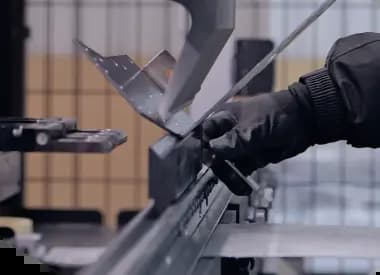 A worker wearing a black protective glove operates an industrial press brake machine to bend a perforated sheet metal part. The metal piece is held firmly between the upper punch and lower die of the machinery.