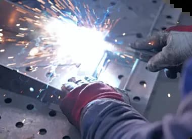 Close-up of a welder in thick protective gloves performing spot welding on a metal workpiece. Bright white light flashes and orange sparks fly outward from the point of contact against a perforated steel welding table.